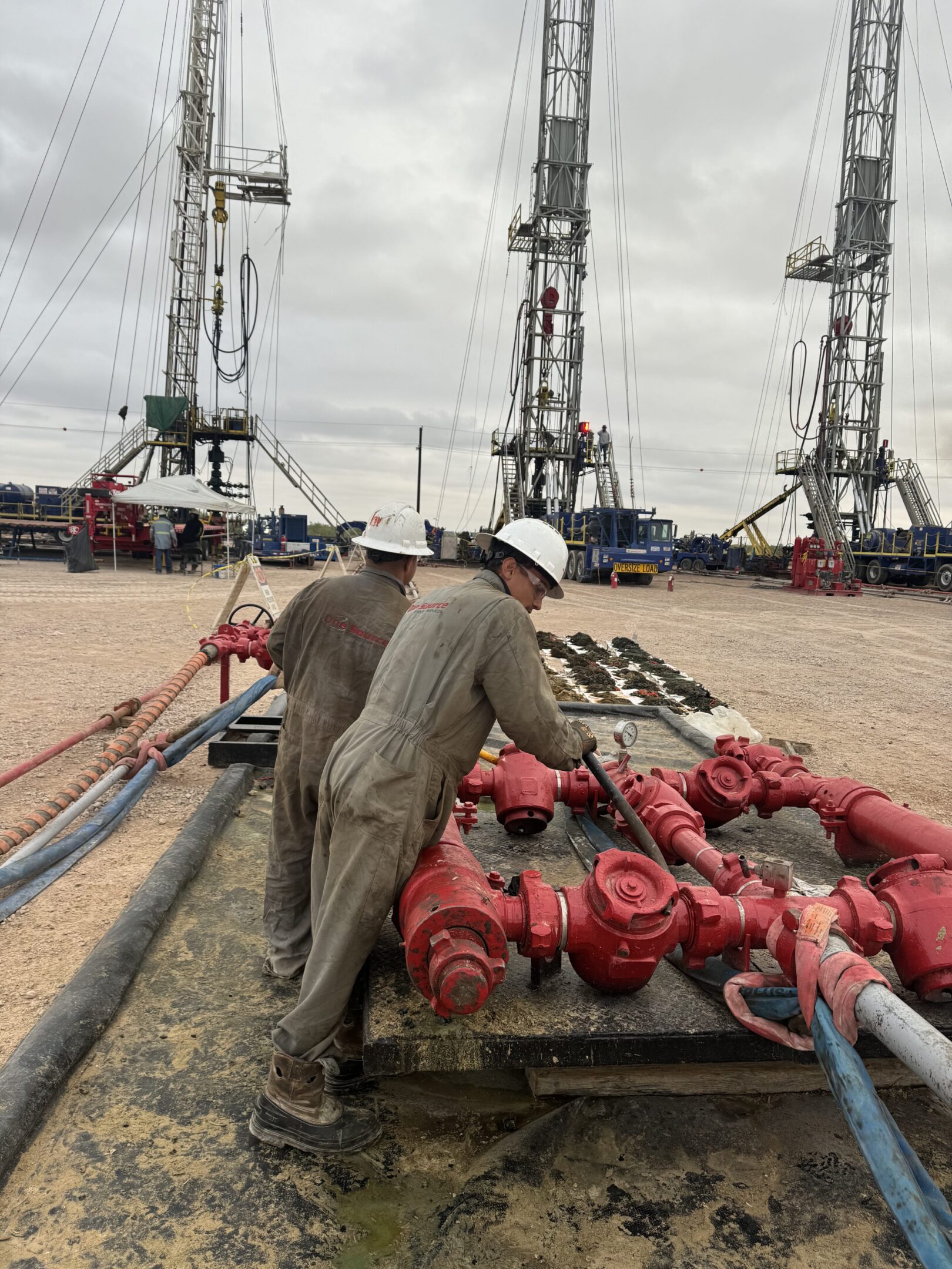 Two workers operating red oil drilling equipment on a cloudy day.